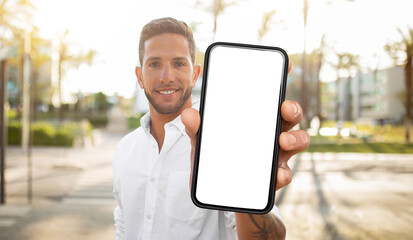 A man in a white shirt stands outdoors, holding a smartphone in front of him with a blank white...