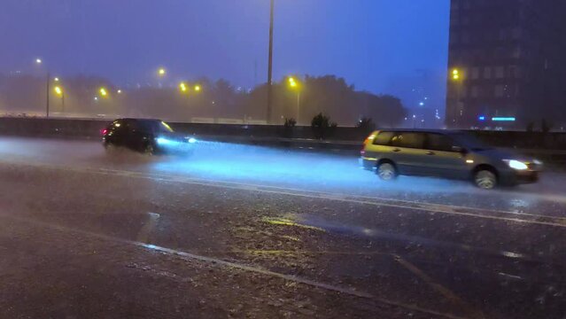 Cars passing through a road flooded by heavy rain. Heavy rain on city street in evening. Car headlights shine through falling raindrops. Summer downpour in city.