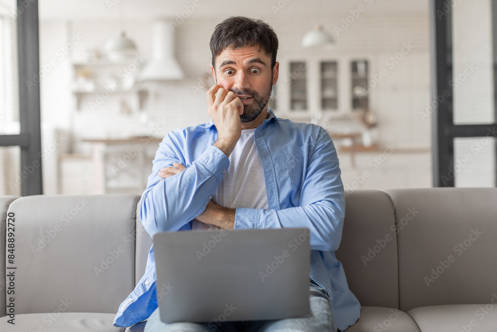 Poster Man sitting on a couch in a home setting. He is looking at his laptop with a worried expression on his face and has his hands clasped together in front of him. He appears to be stressed or anxious.
