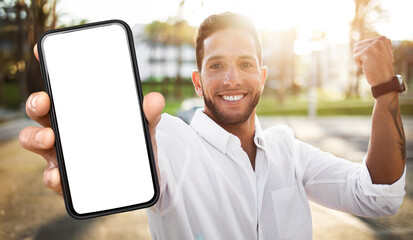 A young man wearing a white shirt stands outdoors in the daylight. He is holding a smartphone with a blank screen in front of him, smiling and looking directly at the camera.