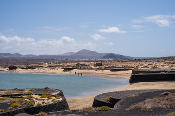 Fototapeta premium Seascape of La Santa Lake. White sand beach and rocks. Mountains in the background. Sky with big white clouds. White houses. Lanzarote, Canary Islands, Spain