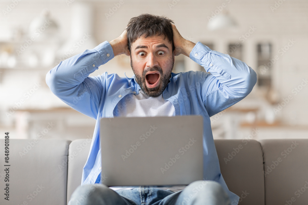 Poster man sitting on a couch, staring in shock at his laptop screen. his hands are on his head, and his mo