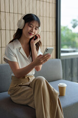 Happy Asian woman listening to music on mobile phone while sitting on sofa at coffee shop Relaxed smiling young woman with headphones in the morning Holiday relaxation concept.