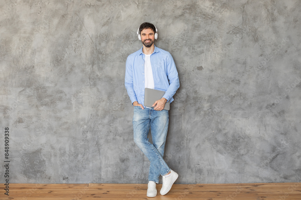 Poster man standing in front of a concrete wall, leaning against it with one hand in his pocket. he is smil