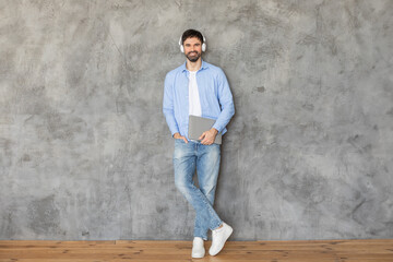 Man standing in front of a concrete wall, leaning against it with one hand in his pocket. He is smiling at the camera and wearing headphones over his ears. He is holding a laptop in his other hand