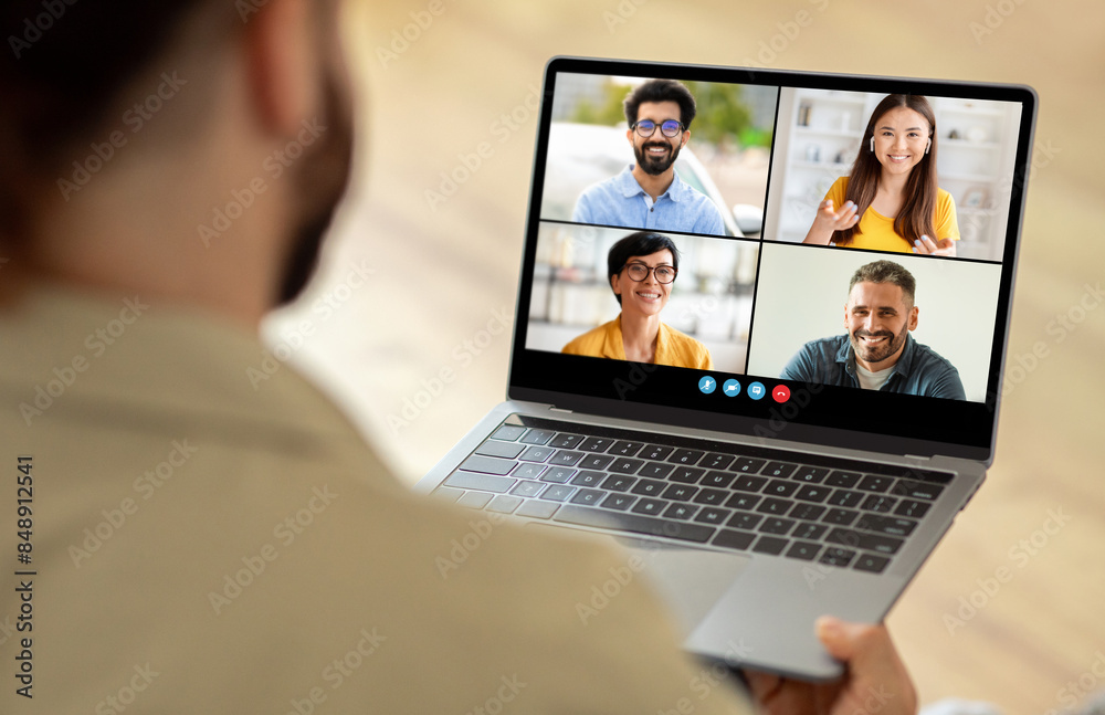 Poster A man sitting at a table is using a laptop, which displays a group of people on the screen. He appears engaged in a virtual meeting or video conference with his colleagues.