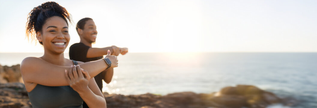 African American woman and man are stretching their arms in front of a beautiful sunset view over the ocean. They are standing on a rocky cliff, with the ocean stretching out behind them, copy space