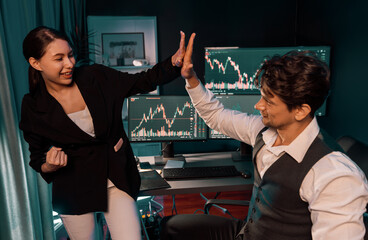 Successful two business traders making hands high five with crashing fist hands for highest profit on pc screens background in real time data, man holding tablet at night light office room. Postulate.