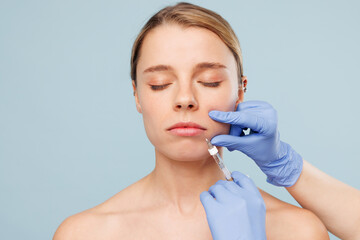 Beautiful calm half naked topless Caucasian young woman with nude make up getting cosmetic injection procedure at clinic isolated on plain blue color background. Skin care healthcare cosmetic concept.