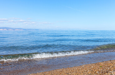 Natural background of clean fine gravel beach with transparent  blue water and ridge of mountains on horizon on sunny clear day. Siberian Baikal Lake. Beautiful landscape of shore of Olkhon Island 