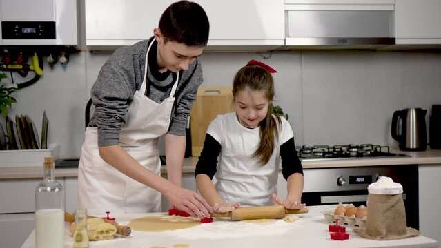 Beautiful girl rolls dough with rolling pin on table. Boy makes cookies with molds by cutting out figures at kitchen. Teenage children brother and sister cooking together. Happy family kids dream
