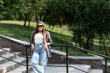 Portrait of a pretty and attractive Caucasian young woman in sunglasses and stylish casual clothes who is happy to pose while walking in the park on the steps on a warm spring day.