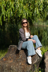 Attractive young woman in sunglasses and stylish casual clothes with pleasure posing sitting on a big stone in the park while walking in the park on a spring day. Relax, freedom.