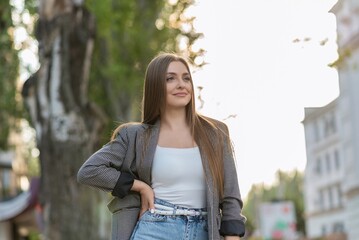 Pretty and attractive Caucasian young woman in stylish casual clothes posing with pleasure while walking in the park on a warm spring day. The concept of freedom, relax, style.