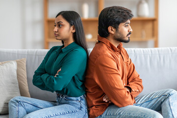 A young Indian couple is sitting back-to-back on a couch, clearly upset and avoiding eye contact....
