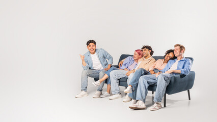 A group of five friends are enjoying each others company, laughing and smiling. The diverse group of young adults are casually dressed in denim and sitting on a couch in a light studio space.