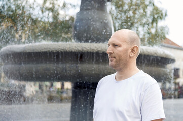 Bald man in white t-shirt standing by fountain looking thoughtful