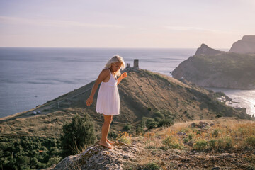 Fototapeta premium A blonde woman stands on a hill overlooking the ocean. She is wearing a white dress and she is enjoying the view.