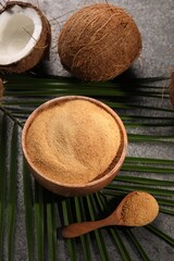 Coconut sugar in bowl, spoon, palm leaves and fruits on dark textured table, above view