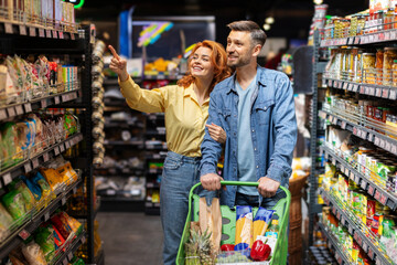 Happy couple with shopping cart walking at supermarket and buying food , woman pointing at shelf with goods, ask husband to buy something