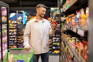 Male customer in supermarket. Man doing grocery shopping alone, taking jar from shelf, choosing...