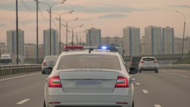 Police patrol car with flashing light drives along highway with city in background.