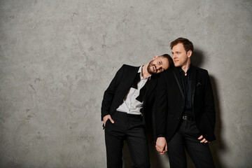 Two men in elegant black suits stand against a grey wall, showing their affection for each other.