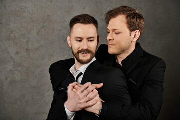Two men in elegant suits embrace against a grey background.