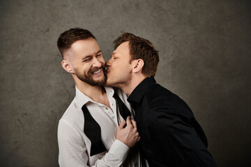 Two men in suits kiss, laughter against grey background.