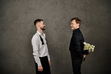 Gay couple in suits hide white flowers behind textured wall.