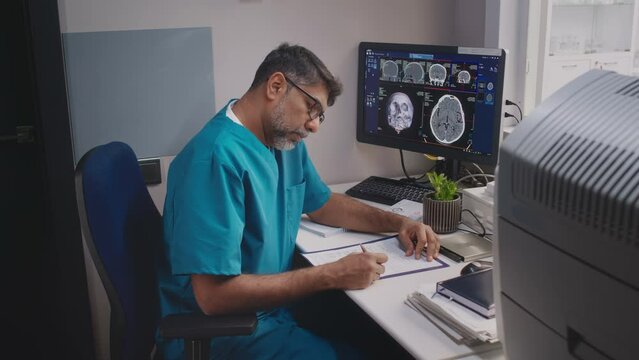 Medium shot of middle-aged male neurologist wearing blue medical uniform sitting at table and writing report after analyzing patients brain MRI scan displaying on computer screen