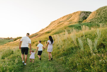 A mom, and dad holding hands baby daughter, son walk in mountains at sunset. Family spending time together on a summer day. Children with parents happiness walking in green grass in field. Back view