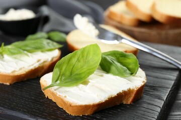 Pieces of bread with cream cheese and basil leaves on black table, closeup