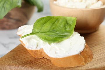 Piece of bread with cream cheese and basil leaf on white table, closeup