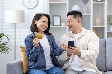 Smiling couple making an online purchase using a phone and credit card while sitting on the sofa at home.