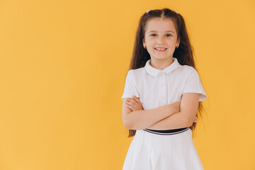 Little girl with dimples on her cheeks smiling and standing with arms crossed wearing a school uniform on a yellow background