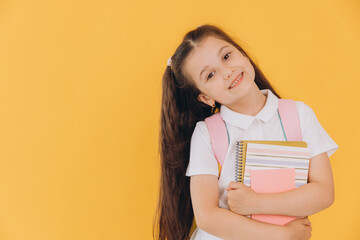 Cute little first grader girl in white school uniform with backpack holding notebooks and books on...