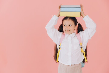 Cute smiling child school girl in uniform with backpack holding books on her head on pink background