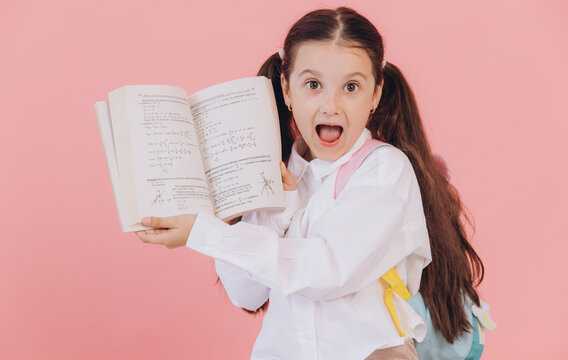 Cute little girl in school uniform with backpack with shocked face and open mouth holding math book and showing equation on pink background