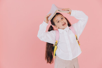 Cute smiling little girl in school uniform with backpack holding open book above head on pink background