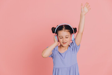 Adorable caucasian kid in blue dress listening to music wearing wireless headset and dancing, isolated over pink background