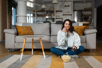 Young happy freshly divorced or recently single woman after toxic relationship breakup sitting alone at home on floor, eating popcorn and watching movie or television in her new apartment she rented.