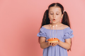 Cute little girl in a blue dress blowing out candles on a donut on her birthday on a pink background