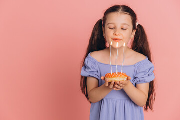 Cute little girl in a blue dress blowing out candles on a donut on her birthday on a pink background