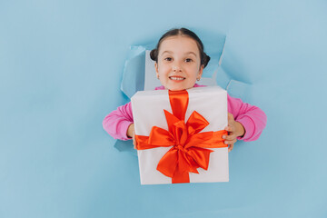 Adorable little girl peeking from ripped paper hole and holding gift. Valentines, Mother day, birthday or celebration concept.