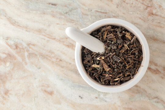 Top-view of Dry Organic Bhringraj (Eclipta prostrata) leaves, in white ceramic mortar and pestle, on a marble background.