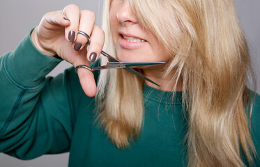 Close up of a blonde Caucasian woman in a green sweater cutting her own hair at home, grey empty...