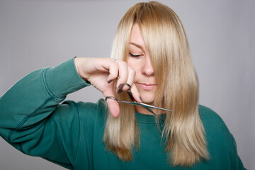 Close up of a blonde Caucasian woman in a green sweater cutting her own hair at home, grey empty...