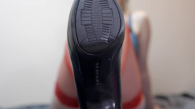 Woman holds out bottom sole of black heel up as she takes off shoes to reveal red panty hose wearing lingerie
