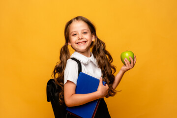 A cute little schoolgirl in a school uniform and with a school bag, books and green apple poses in the studio on a yellow background. Education. Place for text.
